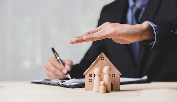 Business man in suit holding hand over small wood house with three small wood people.